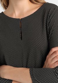 Schwarz gepunktete Bluse mit rundem Ausschnitt, die ein Schlüsselloch-Detail und lange Ärmel aufweist. Der Stoff wirkt glatt und leicht.
