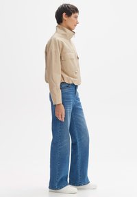 Beige, langärmliges Hemd mit zwei Brusttaschen, kombiniert mit weit geschnittenen blauen Jeans und weißen Sneakers. Glatter Stoff und taillierte Silhouette.