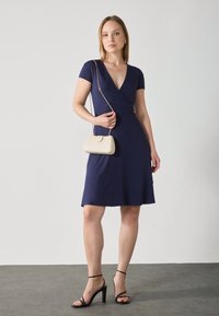 Robe ajustée bleu marine avec des manches courtes et un décolleté en V. Associée à un petit sac à main de couleur crème et des sandales à talons hauts noires.