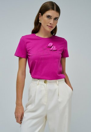 Camiseta de algodón fucsia con mangas cortas, que presenta detalles de aplicación floral y bordado; combinada con pantalones ajustados de color crema.