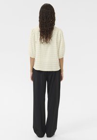 Top testurizzato color beige chiaro con maniche a sbuffo, abbinato a pantaloni larghi neri a righe. La vista posteriore mostra una vestibilità morbida e uno stile casual.