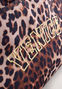 Material cu imprimeu leopard, cu pete amestecate maro și negru. Prezintă brodat cuvântul "VENICE" în galben, cu un font îndrăzneț, geometric.
