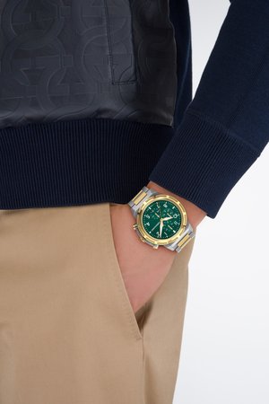 Marineblauwe trui met een gestructureerd zwart paneel, tankleurige broek en een groene wijzerplaat klok met gouden accenten en een zilveren band.