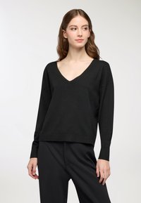 Młoda kobieta w czarnej swetrze z dekoltem w serek i dopasowanych czarnych spodniach stoi na jednolitym jasnym tle, patrząc w lewo.