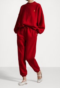 Czerwony sweatshirt i dopasowane dresy wykonane z miękkiego materiału. Posiada okrągły dekolt, elastyczny pasek w talii oraz boczne kieszenie. Szczegółowo zdobione logo.