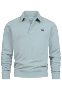 Lyseblå økologisk bomulds polo med lange ærmer, knappet krave og en lille mørk logopatch på venstre bryst.