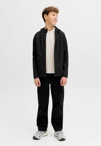 Juodas gobtuvu užsegamas džemperis su priekinėmis kišenėmis, dėvimas ant šviesios spalvos marškinėlių, derinamas su laisvomis juodomis krovinių kelnėmis ir pilkais sportbačiais.