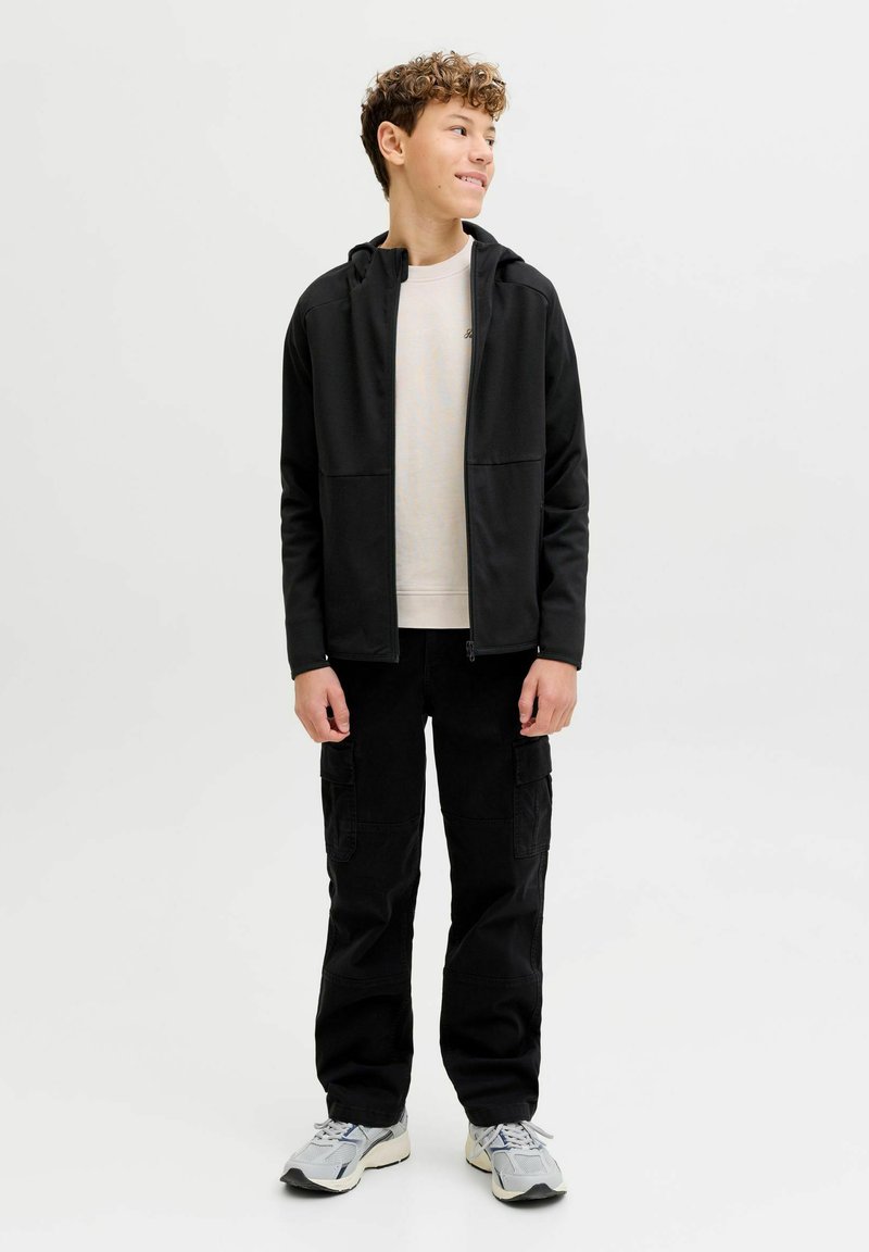 Juodas gobtuvu užsegamas džemperis su priekinėmis kišenėmis, dėvimas ant šviesios spalvos marškinėlių, derinamas su laisvomis juodomis krovinių kelnėmis ir pilkais sportbačiais.