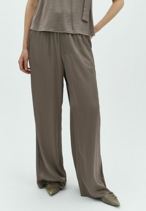 Personne portant un pantalon large et ample couleur taupe avec un haut assorti et des chaussures pointues taupe, debout sur un fond clair uni.