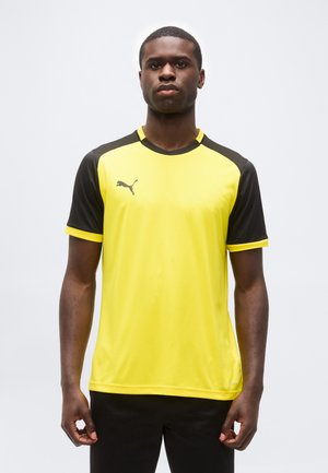 Sárga és fekete Puma sportpólót viselő férfi, karjait lazán tartva, egyszínű világos háttér előtt áll.