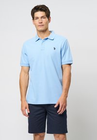 Light blue polo shirt with a classic collar and short sleeves, featuring a small black logo on the chest, paired with dark navy shorts.