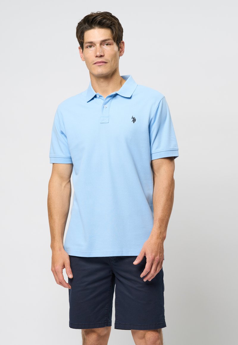 Light blue polo shirt with a classic collar and short sleeves, featuring a small black logo on the chest, paired with dark navy shorts.