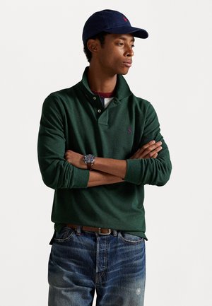 Donkergroene poloshirt met een omgeslagen kraag, twee knopen en een klein paarse logo. Gecombineerd met vervaagde blauwe jeans en een marineblauwe pet.