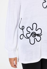 Vitt stickat tröja med en svart broderad blommönster och spiraldetalj. Långa ärmar och ribbade muddmar.