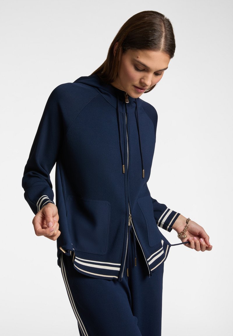 Felpa con zip colore navy, dotata di cappuccio, due tasche frontali, polsini a righe e cordoncini. Tessuto morbido con una texture liscia e dettagli in oro.