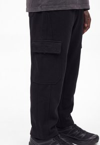 Homme portant un pantalon cargo noir avec une grande poche latérale et des chaussures de sport noires, debout devant un fond blanc uni.