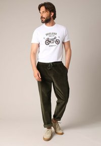 Witte t-shirt met motorfietsafbeelding, gecombineerd met donkergroene corduroy broek met elastische tailleband. Tan schoenen met lichtgekleurde veters.