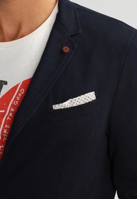 Granatowa marynarka o miękkiej teksturze, z kieszenią na piersi, w której znajduje się biała chusteczka z niebieskimi wzorami. Prosty detal guzika na klapie.