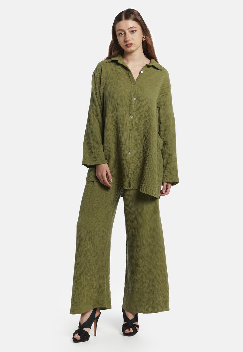Olivengrön, texturerad, knappad skjorta med långa ärmar, i kombination med vida byxor. Modellen bär svarta sandaler med öppen tå.
