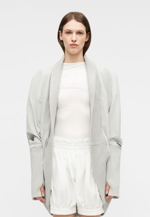 Persona con un cárdigan extragrande de color gris claro, camiseta blanca de manga larga y pantalones cortos blancos con cordón, de pie frente a un fondo liso.