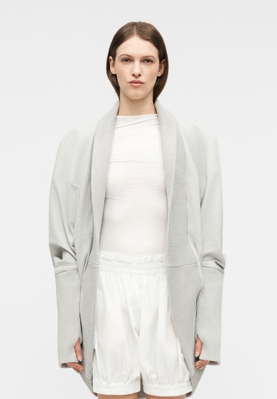 Personne portant un cardigan oversize gris clair, un haut à manches longues blanc et un short à cordon blanc, debout devant un fond uni.