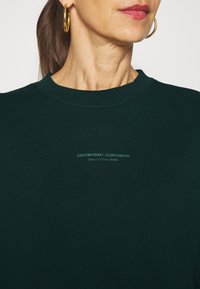 Donna che indossa una maglietta verde scuro a collo rotondo con del testo in piccolo "CONTEMPORARY_SCANDINAVIAN Marc O’Polo DENIM" e orecchini a cerchio dorati.