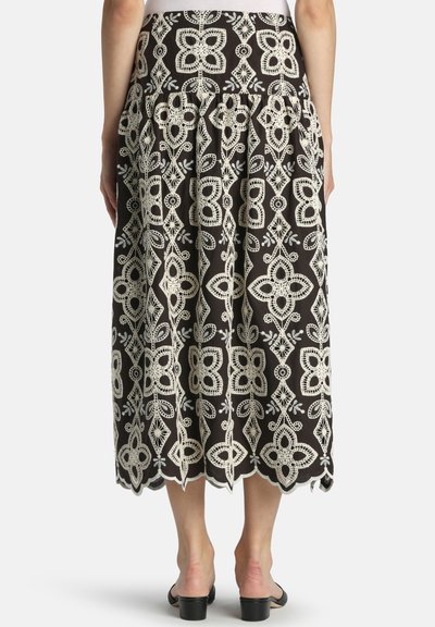 Zwarte midirok met ingewikkeld wit bloemenkantborduursel en gezoomde zoom, gedragen door een persoon die staat met voeten in zwarte sandalen met hak.