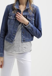Žena oblečená v modré džínové bundě přes šedé tričko a bílé kalhoty, s rukou upravující límec bundy na jednolitém pozadí.