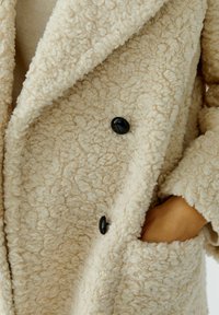 Beige textured coat made of plush fabric, featuring a double-breasted design and black buttons. Includes side pockets for utility.