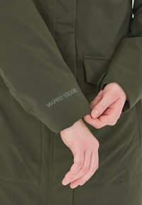 Groene waterdichte jas met een gladde textuur en een zichtbare "W-PRO 10000" print op de mouw. De handen zijn bezig met het verstellen van de manchet.
