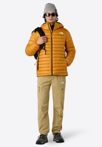 Žlutá nadýchaná bunda s horizontálním prošíváním, černými zipy, doplněná béžovými kalhotami a černými botami. Model nosí sluneční brýle a šedou čepici.