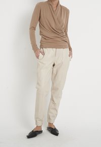 Sandfarbene Langarmbluse mit Drapierung, kombiniert mit beigen, schmal zulaufenden Hosen mit elastischen Bündchen und schwarzen Slippers.