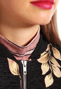 Černá bomber bunda s propracovanou růžovou květinovou výšivkou a stříbrným zipem. Pod ní světle růžová košile s límečkem a jemným proužkováním.