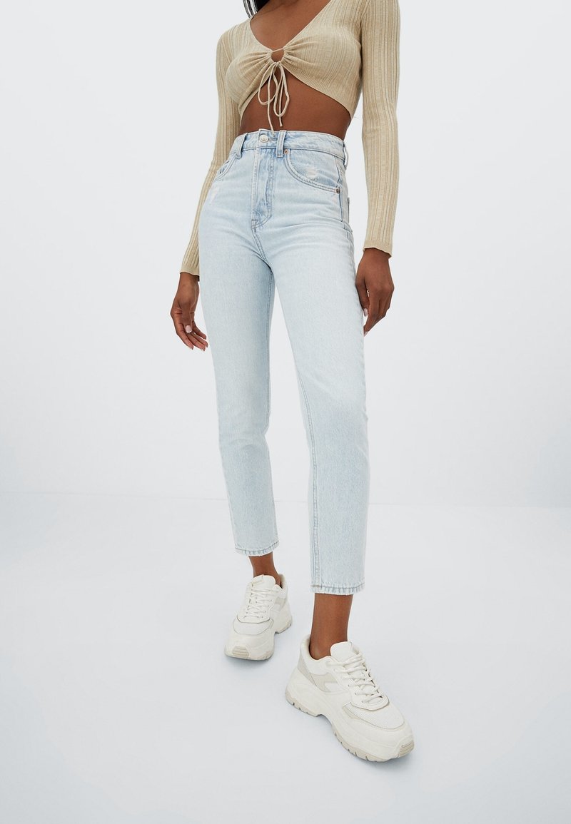 Femme portant un jean taille haute bleu clair, un top beige froncé court à manches longues et des baskets blanches épaisses, debout devant un fond blanc.