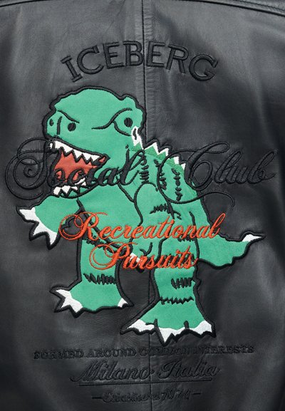 Πράσινος καρτουνίστικος δεινόσαυρος με αιχμηρά δόντια κεντημένος σε μαύρο δέρμα, με κείμενα "ICEBERG," "Social Club," και "Recreational Pursuits" σε διακοσμητικές γραμματοσειρές.