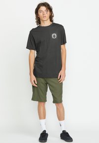 Sort bomulds T-shirt med grafisk lomme på forsiden, parret med olivengrønne ternede shorts, hvide sokker og sorte sko.