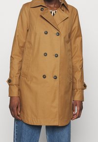 Žena oblečená v béžovém dvouřadovém trenčkotu s knoflíkovými poutky na rukávech a modrými džíny, stojící na jednobarevném pozadí.