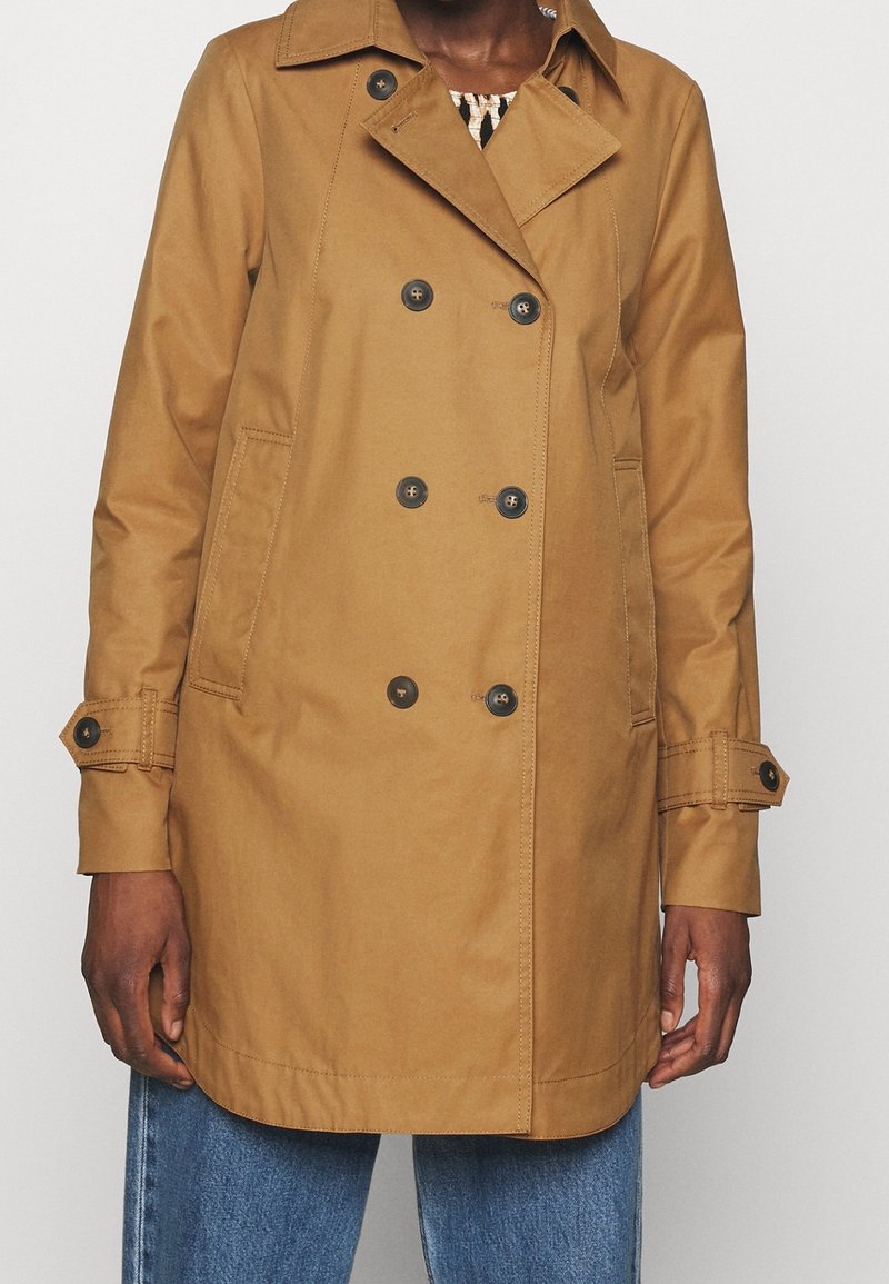 Žena oblečená v béžovém dvouřadovém trenčkotu s knoflíkovými poutky na rukávech a modrými džíny, stojící na jednobarevném pozadí.