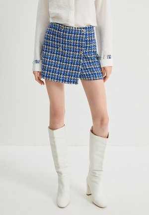 Pantaloni shorts in tweed a scacchi blu e bianchi con bottoni dorati, dal taglio sartoriale. Abbinati a stivali bianchi con tacco al ginocchio.