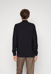 Svart strikket cardigan med høy krage, ribbestrikket hem og mansjetter, lange ermer og en glatt tekstur. Brukt over brune mønstrede bukser.