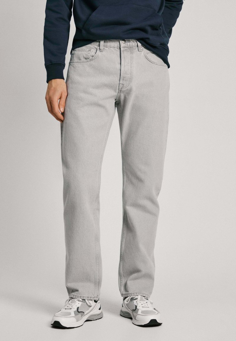 Gai pelēki džinsi ar taisno kāju dizainu, ar piecām kabatām un redzamu pogu un rāvējslēdzēja aizdari. Valkāti ar baltām sporta apaviem.