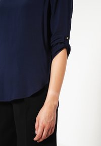 Mörkblå blus med uppvikta ärmar, mjuk textur och böjd fåll. Guldknappdetalj på ärmen ger en subtil accent.