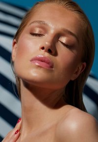 Close-up of a face with smooth, glowing skin, light pink lips, and bronze eyeshadow; background features bold blue and striped patterns.