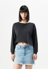 Woman with straight dark hair wears a loose black cropped long-sleeve top and a light blue frayed denim mini skirt, standing against white background.