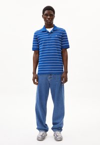 Blau gestreiftes Poloshirt mit Kragen und kurzen Ärmeln, kombiniert mit lockeren blauen Jeans und grauen Sneakern. Modell steht vor einem weißen Hintergrund.