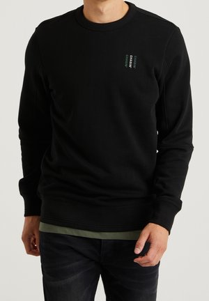 Sudadera negra con cuello y puños acanalados. Presenta texto verde en el pecho. Textura suave, corte relajado, adecuada para uso casual.