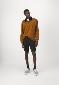 Polo a maniche lunghe marrone con colletto nero, abbinato a pantaloni corti neri e sneaker grigie. Il polo presenta un logo sul petto.