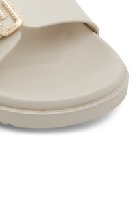 Sandale à glisser beige clair avec une texture lisse et une large sangle ornée d'une boucle rectangulaire dorée. Semelle basse.