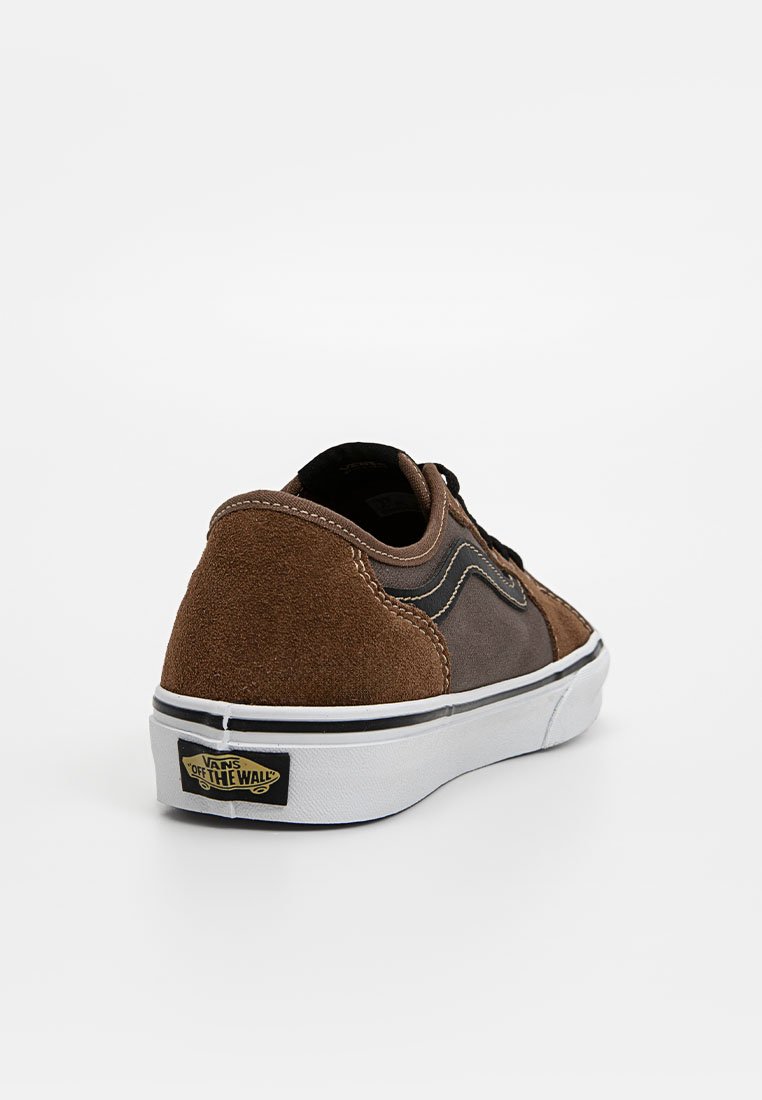 Zapatilla baja de ante marrón con suela de goma blanca, acentos negros en contraste y un logo de Vans en el talón. Parte superior texturizada y detalles de costura.