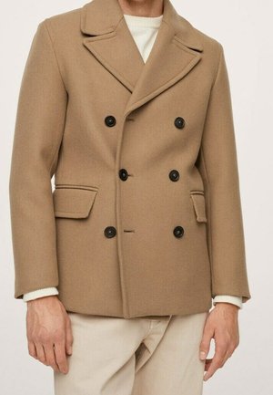 Uomo che indossa un cappotto doppiopetto color cammello con bottoni neri sopra un maglione bianco e pantaloni beige.